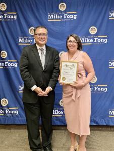 Erica Luna Gonzales stands with Assemblymember Mike Fong holding an Assembly certificate of recognition in front of an Assemblymember Mike Fong District 49 backdrop