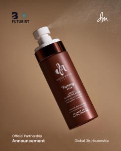 A dramatic angled photograph of a floating brown bottle of Danessa Myricks Liquid Blurring Balm, against a warm brown gradient background. A fine mist is being sprayed from the white nozzle.