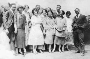 A dozen Black people pose for a photo. They seem comfortable together and friendly. The eight women are dressed per mid-1920s daytime fashion in dropped-waist dresses with hems well below the knee and low-heeled shoes. The four men are all dressed in dark