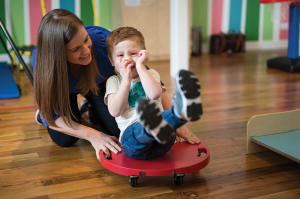 A woman in a blue shirt is smiling at a young boy sitting on a scooter while the woman is pushing him.