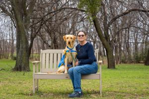 A woman sits on a bench beside a yellow labrador/golden retriever dog wearing a service dog vest.