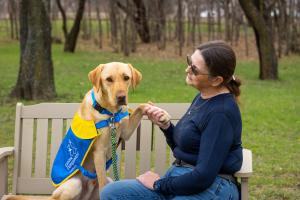 A woman sits on a bench beside a yellow labrador/golden retriever dog wearing a service dog vest. The woman is holding the dog's front paw in her hand.