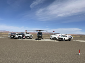 Helicopter centered on a runway surrounded by multiple fuel trucks, with mesas and open sky in the background.