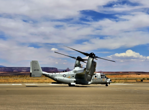 Military tiltrotor aircraft on a runway with rotors angled upward, set against a desert landscape and cloudy sky.