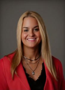 Woman with blonde hair and wearing a red jacket smiling in a professional picture after being awarded the Distinguished Cheetah Alum Award
