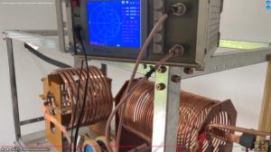 A Vector Network Analyzer displaying a blue Smith chart sits atop a metal frame. Coaxial cables connect the device to large, copper-wound inductor coils housed within the rack below.