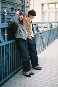 Deux personnes en jeans en denim brut à revers, appuyées contre une structure métallique dans un environnement urbain.