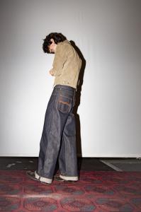 Personne portant un jean en denim brut à coupe large avec ourlet retroussé et veste beige, debout sur un tapis dans un espace intérieur minimaliste.