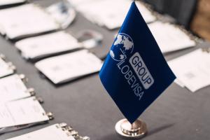 A close-up shot of a blue desktop flag featuring the Globevisa Group logo and a white globe icon, set against a background of blurred professional event name tags and lanyards on a table.