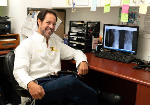 Photo of Dr. Gregg Rubinstein in a white shirt with the 57th Street chiropractic logo in green and yellow, seated at a desk.