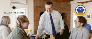 Waiter serving drinks to a group of people seated at a table in a cozy restaurant with Black Star Farms and Certified Autism Center logos.