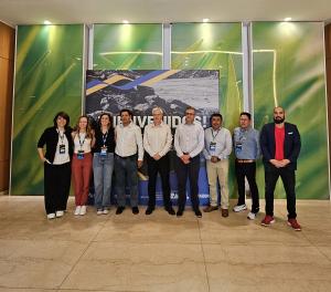Group of ten people standing side by side in an indoor setting. In the background, there is a panel displaying the word “Welcome” along with visual elements in green and neutral tones, suggesting an institutional event related to the mining sector. The in