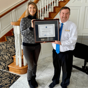 Two people holding a framed certificate together, standing in a room with a staircase and marble floor.