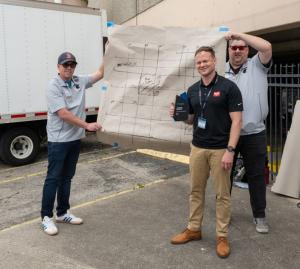 Two of them are holding a large paper sheet with blue tape on the corners and hand drawn markings made with a marker representing concrete scan results. One person is standing in front of the paper and the other two people holding small black and blue plaque.