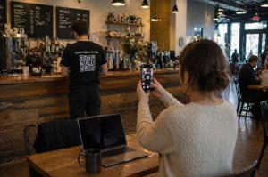 Customer scanning a Dont Click On Me QR code shirt from a distance in a café setting while a worker wears the shirt, demonstrating interactive apparel in real-world use