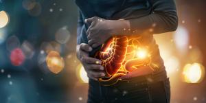 A person in dark clothing holds their abdomen in discomfort while a glowing, semi-transparent illustration of the digestive system overlays their torso, highlighting the intestines with warm orange and yellow light against a softly blurred background.