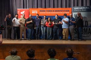A group of student winners and adult industry representatives stand on a stage in front of Project MFG and General Dynamics Electric Boat banners, holding awards and posing for a photo while an audience watches from below.