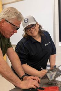A woman wearing a Project MFG hat and General Dynamics Electric Boat shirt smiles while reviewing a welded metal piece with a colleague at a table, assessing the work during a skills competition.