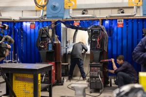 Inside a workshop, a student works in a welding booth surrounded by blue protective curtains, while another person crouches beside a welding machine adjusting controls, with equipment and tools visible throughout the space.