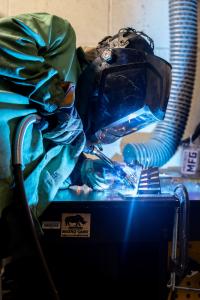 Close-up of a welder wearing a protective helmet and gloves, using a welding torch on a metal piece as bright sparks and blue light illuminate the workspace.