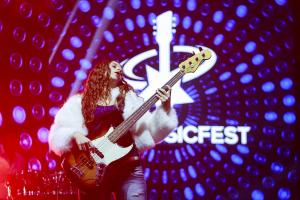 Female musician playing bass guitar on stage at Tour Music Fest, with red and blue lighting and event logo in the background.