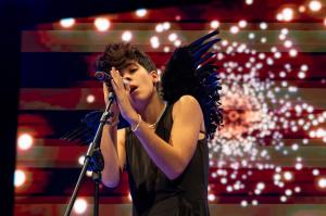 Singer performing on stage at Tour Music Fest with microphone, wearing black outfit with feathered wings and colorful light effects in the background.