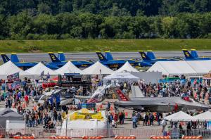 Crowds explore aircraft displays and exhibits at an Air Dot Show event, with U.S. Navy Blue Angels jets visible in the background.