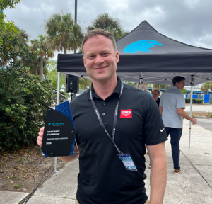 GPRS Chief Services Officer Jamie Althauser wearing a black polo with the red GPRS logo and a lanyard around his neck. He is holding a blue and black plaque labeled “Concrete Champion Proceq GPR Tagging Competition 2026.” He is standing outdoors in front