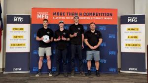 Four members of the Thaddeus Stevens College of Technology team stand in front of a Project MFG backdrop, holding machined parts and components, indicating their advancement to the national championship finals.