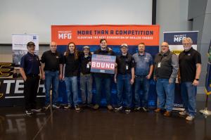 A group of students and instructors stand in front of a Project MFG backdrop, with one person holding a “National Championship Bound” sign, indicating the team has advanced as one of four finalists in the competition.