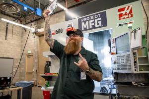 A competitor from Great Basin College stands in a machine shop in front of a CNC machine, raising a freshly machined metal part while making a celebratory hand gesture.