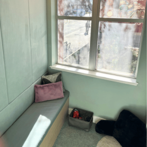 Cozy corner with a window, light green walls, a cushioned bench with pillows, and a small storage bin on a carpeted floor.