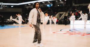 Ashwin Gane walking across an NBA court mid-performance during halftime with arena lights and audience in the background.