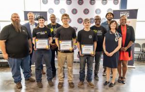 A group of competition winners and organizers pose indoors in front of Project MFG banners. Three young participants at the center hold certificates of achievement, while other adults stand beside and behind them, smiling. The setting appears to be an awa