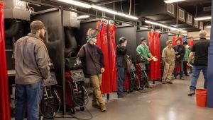 A row of welding booths inside a workshop, each equipped with welding machines and red safety curtains. Several young participants wearing protective gear such as welding jackets, gloves, and caps stand at their stations while listening to an instructor a