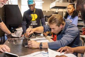 A man leans over a metal worktable, using a precision measuring tool to inspect a welded piece while several observers stand nearby. Papers with diagrams and notes are spread across the table, indicating the scoring process during a welding competition.