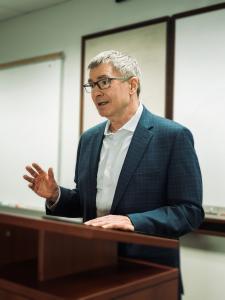 Middle-aged man with glasses standing behind wood podium, one hand lifted as he engages the audience in story.