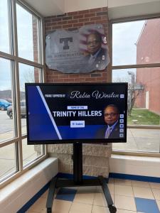 Interactive touchscreen display on a mobile stand at Trinity High School showing a searchable digital archive of yearbooks and alumni history, with a donor plaque mounted above.