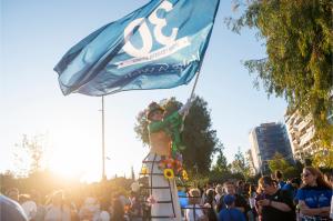 A stilt-walker smiles above a crowd against a blue sky waving a blue flag