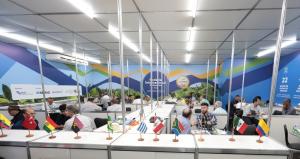 Indoor environment organized in a business matchmaking format, with multiple tables arranged in rows and separated by metal dividers. At each table, small groups of people sit face to face, talking and negotiating. Most are adult men dressed in business c