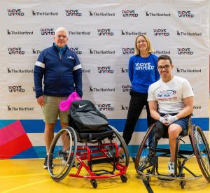 Three individuals pose in front of a banner with adaptive sports equipment in front of them