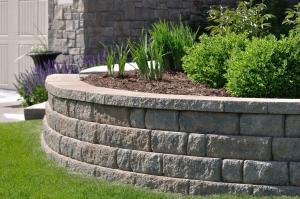 Close-up of a tiered, curved stone block retaining wall holding a garden bed with mulch, green shrubs, and purple flowers.