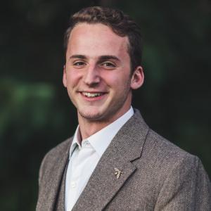 A professional portrait of Aaron Cherry wearing a textured grey blazer with a gold bee lapel pin.