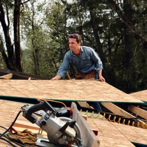 Aaron Cherry in a tool belt lifting a sheet of plywood while working on a roof alongside professional power tools.