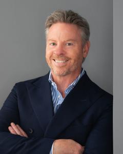 Professional portrait of Brett McAllen, CEO of @WORKSPACES. A middle-aged man with short, styled grey-brown hair and light facial hair, wearing a navy double-breasted blazer over a blue-and-white striped shirt. He is smiling warmly with his arms crossed, 