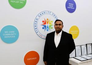 A portrait of Clayburn Figredo in a black suit standing against a white wall featuring the 'Figredo Charity Foundation' logo and colorful partner discs, including 'World Vision Australia' and 'Compassion Australia'.