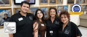 Four people posing in front of framed artwork in a gallery, three wearing matching black shirts with logos.