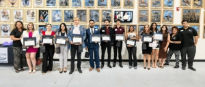 Group of diverse people standing indoors holding certificates in front of a wall covered with framed artwork and photos.