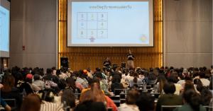 Tagumporn "Gut" Thansiphop and a co-presenter on stage teaching the Maha Taksa astrology system to a large, seated audience in a conference hall. A large projection screen behind them displays a structured grid with numbers and Thai script detailing the a