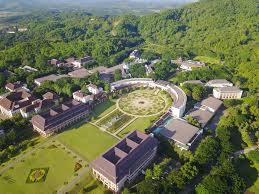 An aerial view of the lush, green campus of Mae Fah Luang University in Chiang Rai, Thailand, featuring modern semi-circular and rectangular buildings built into a forested hillside surrounding a central landscaped garden.